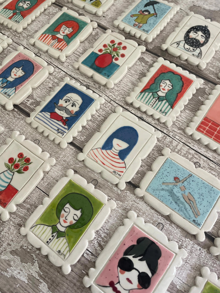 Hand-painted ceramic Mini Masterpieces by Sister Sister — tiny framed artworks featuring colourful portraits, flowers, and whimsical designs on a rustic wooden background