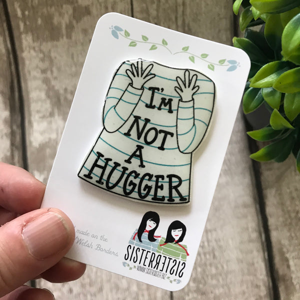 Photo shows a Pin  shaped as a white long sleeve top with turquoise stripes and hand and arms held up in front of chest and the words "I'm Not A Hugger written across the top"  on a white backing card being held against a white wooden background