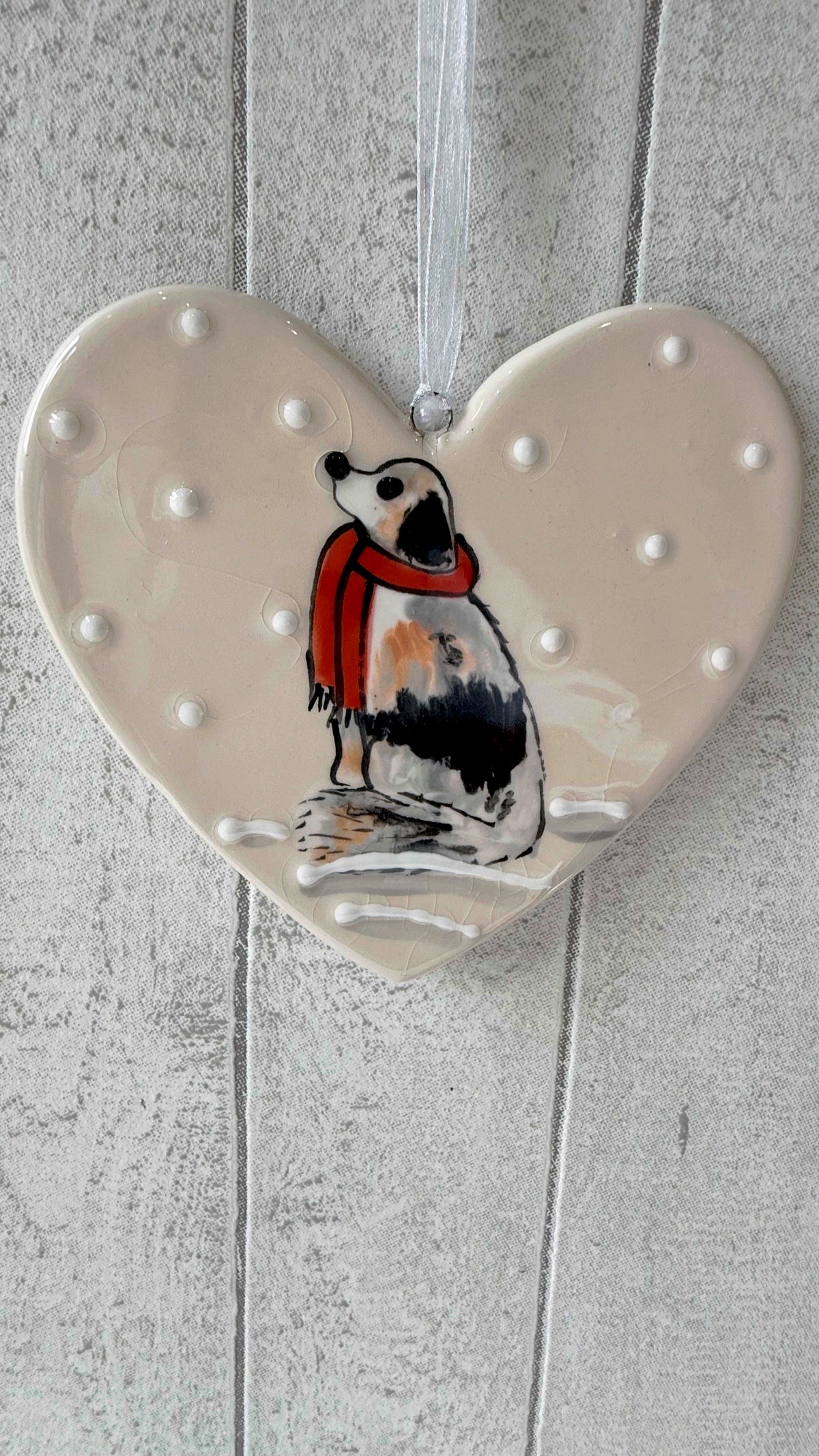 A heart-shaped ceramic ornament featuring a painted dog wearing a red scarf, hanging against a light gray wood plank background.