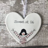 White ceramic heart hand painted with "Sweet at 16".  Dolly in grape top, arms up in celebration, rosy cheeks, heart-shaped lips.  Heart hung with white organza ribbon on a painted white wooden background