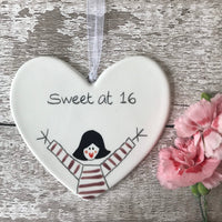White hand painted ceramic heart with "Sweet at 16" text. Adorable dolly in grape Breton top, arms raised, rosy cheeks, heart-shaped lips. Heart is hung with a White Organza Ribbon on a painted white wood background with a pink carnation
